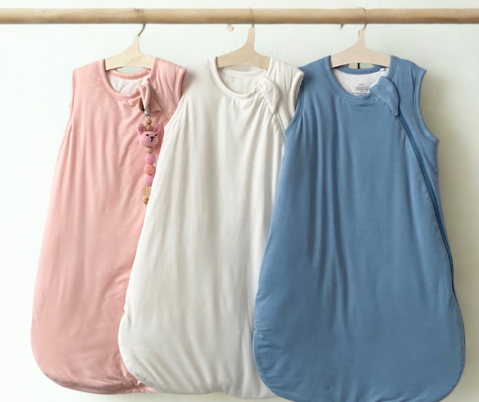 Three baby sleep sacks in pink, white, and blue hanging on a rack against a white background.